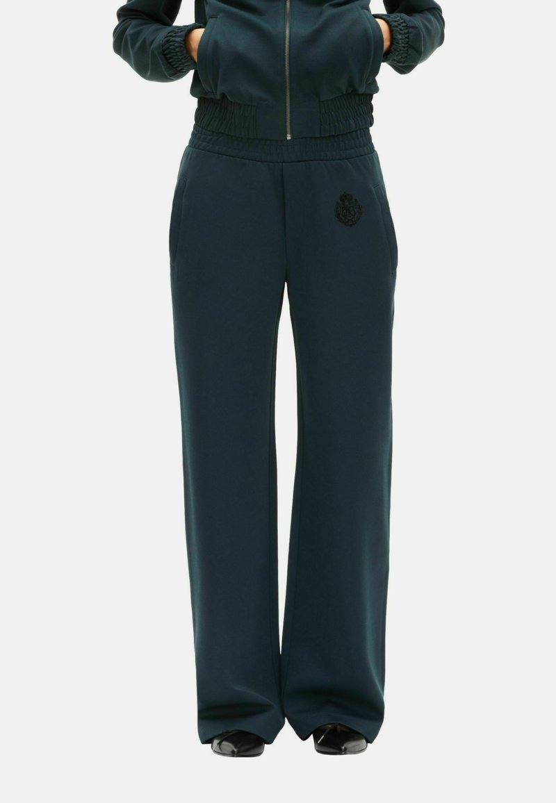 Mørkegrønne joggebukser med en afslappet pasform, elastisk talje, sidelommer og et struktureret emblem nær venstre hofte. Blødt stof.