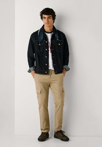 Mörk denimjacka med en ljus denimkrage, vit t-shirt med röd text, beiga cargo-byxor och mörkbruna sneakers med gröna sulor.