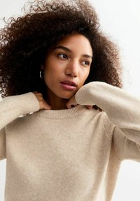 Pull en tricot beige avec un col rond, des poignets côtelés et une texture douce, porté par une personne aux cheveux bouclés, tirant sur le col.