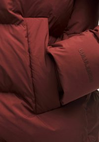 Bordeaux farvet jakke med quiltet design og en glat tekstur. Har et hævet brandlogo på ærmet. Isoleret for varme.