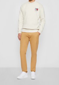 Ljusbeige sweatshirt med rundad hals och liten logotyp på bröstet, matchad med tanfärgade chinos och vita sneakers, allt visat på en enkel bakgrund.