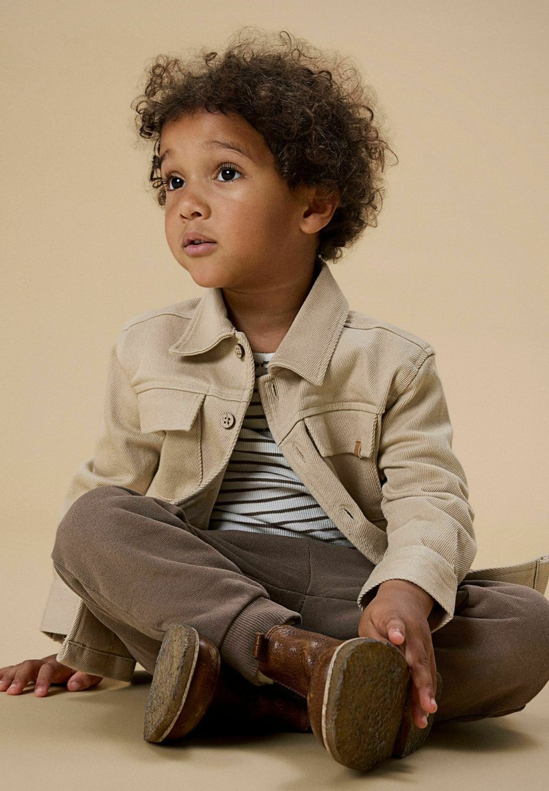 Bézs kordbársony dzseki két mellzsebbel, csíkos ing, barna melegítőnadrág és barna bokacsizma. Gyerek törökülésben ül.