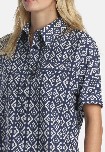 Moteris dėvi tamsiai mėlyną marškinių tipo palaidinę su baltais geometriniais rombo raštais ir krūtinės kišene, matosi šviesūs plaukai, šviesi fonas.