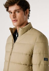 Chaqueta acolchada de color beige claro con cuello alto, textura acolchada y cremallera frontal. Presenta un parche de marca en la manga izquierda.