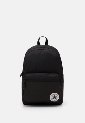 Sort stof rygsæk med en afrundet form, frontlomme med lynlås og et cirkulært Converse All Star-logo på den nederste del.