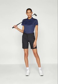 Donna in piedi con una mazza da golf sulle spalle, indossando una polo blu marino, pantaloni corti neri, calzini bianchi e sneakers bianche su uno sfondo neutro.