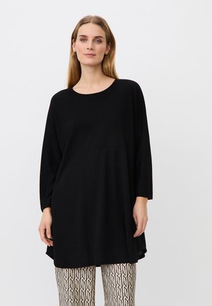 Vrouw staat, draagt een losse zwarte tuniek met lange mouwen en een crèmekleurige en zwarte broek met patroon, naar voren gericht tegen een effen achtergrond.