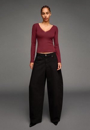 Vrouw die tegen een grijze achtergrond staat, gekleed in een nauwsluitende bordeauxrode top met lange mouwen en losse zwarte wijde broek met puntige zwarte schoenen.