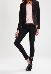 Blazer negro ajustado sobre una blusa rosa clara, combinado con jeans skinny negros y zapatos negros. El blazer tiene un solapa de satén y un diseño entallado.