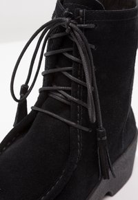 Black suede ankle boot with rounded toe, textured surface, and flat black laces featuring a tassel detail. Low, solid outsole.