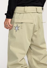 Licht khaki cargobroek met een sterpatch, kleppen op de achterzakken en een gladde textuur. Ritsdetails op de zijnaad.