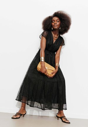 Femme aux cheveux afro portant une longue robe noire en dentelle, des bijoux en or, des sandales à talons noires, et tenant une pochette tissée beige.