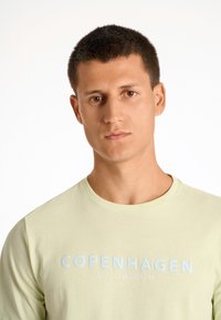 Camiseta de manga larga verde clara con cuello redondo, que presenta un texto azul claro "COPENHAGUE LINDBERGH" centrado en el pecho. Textura suave.