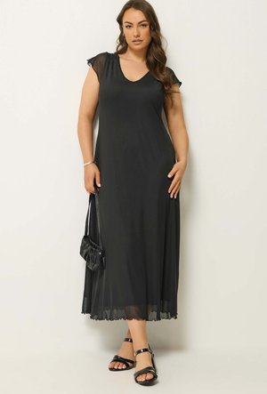 Femme portant une robe longue noire sans manches avec des manches courtes transparentes, tenant un sac à main noir et portant des sandales noires à lanières.