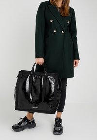 Czarna, połyskująca torba na ramię z podwójnymi uchwytami, z logo Calvin Klein; zestawiona z ciemnozielonym płaszczem i sportowym obuwiem.