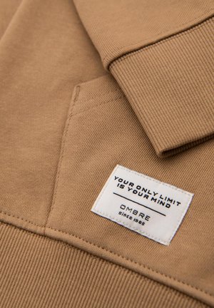 Beige Sweatshirt mit gerippten Bündchen und einer Brusttasche. Auf der Vorderseite befindet sich ein weißes Etikett mit schwarzem Text: "DEINE EINZIGE GRANZE IST DEIN GEIST, OMBRE seit 1985."