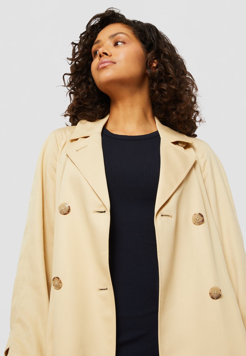 Impermeabile beige con design doppiopetto, grandi bottoni e colletto, indossato su una maglia a coste blu navy. Tessuto morbido, vestibilità rilassata.