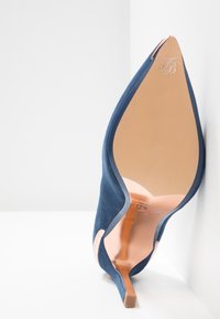 Chaussure à talon haut en daim bleu à bout pointu inclinée pour montrer la semelle lisse couleur beige et le talon aiguille fin sur fond blanc.