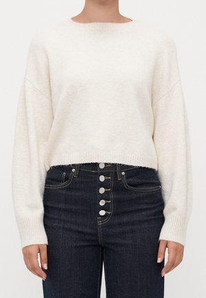 Vrouw draagt een witte, zachte trui met lange mouwen, ingestopt in een donkerblauwe hoge taille jeans met vijf zichtbare zilveren knopen.