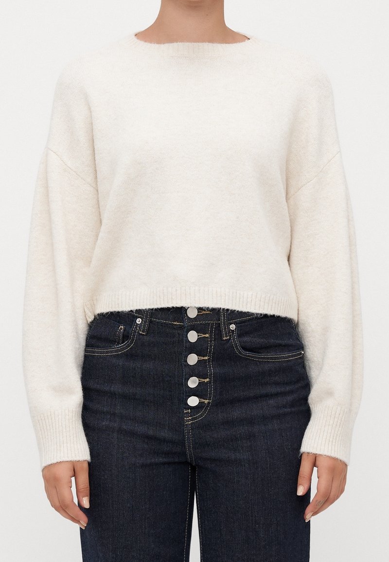 Femme portant un pull blanc à manches longues en tissu duveteux, rentré dans un jean bleu foncé taille haute avec cinq boutons argentés visibles.