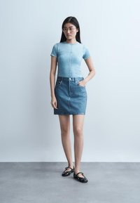 Νεαρή γυναίκα με μακριά σκουρόχρωμα μαλλιά και γυαλιά φοράει ένα ανοιχτό γαλάζιο εφαρμοστό κοντομάνικο τοπ, φούστα από τζιν και μαύρες μπαλαρίνες, στέκεται με το ένα χέρι στην τσέπη.