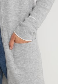 Ljusgrå stickad cardigan med en framficka och kontrasterande vit kant på ärmarna. Mjuk textur och avslappnad passform synlig.