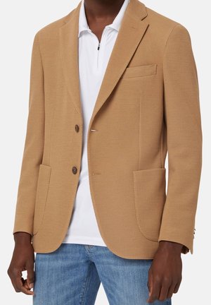 Mand iført en tan single-breasted blazer over en hvid zip-up polo shirt og blå jeans, stående med armene afslappet ned langs siderne.