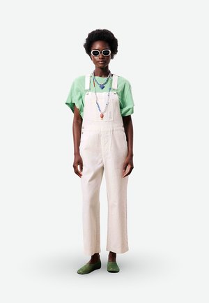 Femme avec afro courte portant une salopette blanche, un t-shirt vert à manches courtes, des chaussures vertes, des lunettes de soleil blanches et des colliers colorés en perles, debout.