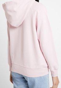 Sudadera rosa claro hecha de tela suave, con capucha, puños y dobladillo acanalados. La parte trasera tiene un ajuste holgado con un dobladillo curvado.