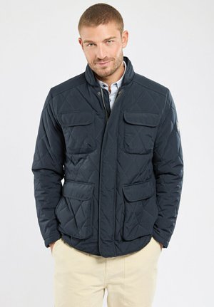 Homme portant une veste matelassée bleu marine avec quatre poches à rabat, un pantalon beige et une chemise à col bleu clair, debout devant un fond uni.