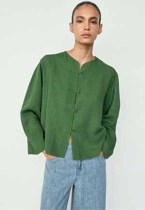 Person trägt ein grünes, geknöpftes Langarmshirt und hellblaue Jeans und steht vor einem einfarbigen hellen Hintergrund.