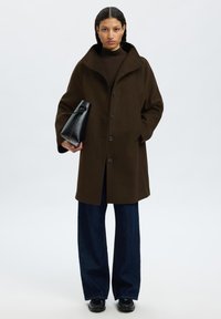 Manteau en mélange de laine marron avec un col haut, fermeture à boutons devant et poches latérales ; porté sur un jean large noir et accompagné d'un sac noir.