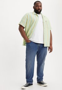 Camisa de manga corta a rayas verdes y amarillas sobre una camiseta blanca, combinada con jeans azules y zapatillas negras con acentos blancos.