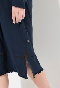 Granatowa sukienka z miękkiego materiału, z falbaniastymi brzegami, bocznymi rozcięciami oraz dekoracyjnym logo na dole dla dodatkowego akcentu.