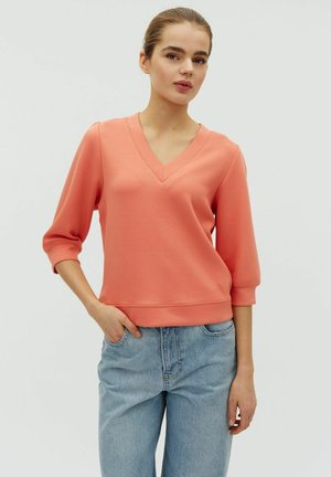 Mlada ženska, oblečena v koralni V-izrez z rokavi do komolcev in svetlo modre kavbojke, z eno roko v žepu, stoji pred enostavnim ozadjem.