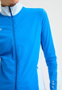 Chaqueta deportiva azul con cuello alto, paneles laterales blancos, cremallera completa y marca visible. Textura de tela suave y ligera.