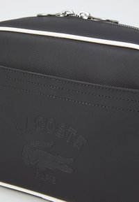Svart texturerad zip väsk med präglad Lacoste-logotyp och krokodildesign, med vit accentkant och en framficka.