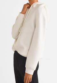 Sudadera blanca de tela suave, con capucha ajustable, puños de canalé y corte holgado, combinada con pantalones negros.