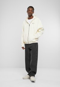 Kremfarget zip-up hettegenser med kengurulommer, kombinert med mørkegrå bukser og beige joggesko, poserer mot en enkel hvit bakgrunn.