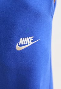 Kék Nike anyag sima textúrával, fehér hímzett logóval. Nagyítás távoli látványa részletes varrást és logótervet mutat.