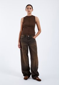 Haut sans manches marron à côtes associé à un jean marron foncé à coupe décontractée, avec poches avant et une texture légèrement délavée. Ballerines marron douces.
