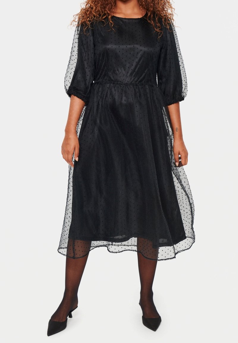 Femme portant une robe noire mi-longue avec un voile transparent à pois et des manches trois-quarts bouffantes, associée à des collants transparents noirs et des talons noirs.
