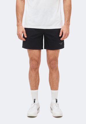 Mężczyzna noszący czarne sportowe spodenki, białe skarpety i białe buty sportowe, stojący z rękami swobodnie opuszczonymi wzdłuż ciała.