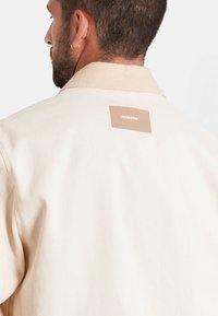 Beige jas van een gestructureerde stof, met een geribbelde kraag en een rechthoekig leren label met "SIXTH JUNE" in reliëf.