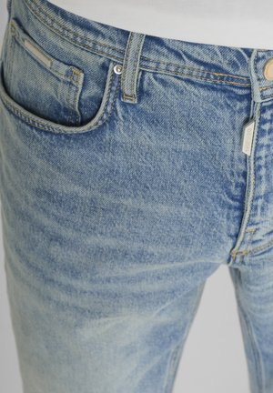 Gros plan sur un jean en denim bleu clair montrant la poche avant, la passants de ceinture, le bouton et les détails de couture visibles.