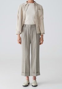 Femme portant une chemise en velours côtelé beige sur un t-shirt blanc, un pantalon large taille haute gris, et des mocassins crème, debout devant un fond uni.
