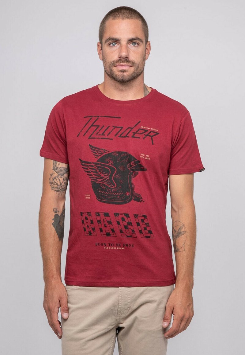 T-shirt en coton rouge avec un design graphique noir comprenant le texte "Thunder" et "RACE", ainsi qu'une illustration de casque ailé. Manches courtes.