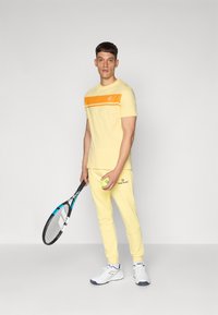 Ensemble de sport jaune comprenant un haut à manches courtes avec une bande orange et un logo, associé à un pantalon assorti. Tient une raquette de tennis et une balle.