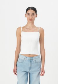 Biely rebrovaný crop top s tenkými ramienkami. Kombinovaný s svetlo modrými džínsy s vysokým pásom, umiestnený na neutrálne pozadí.