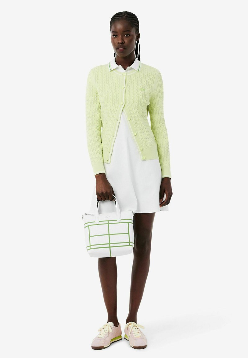 Woman with braided hair wearing a light green cardigan over a white dress, holding a white and green checkered handbag and pink sneakers.